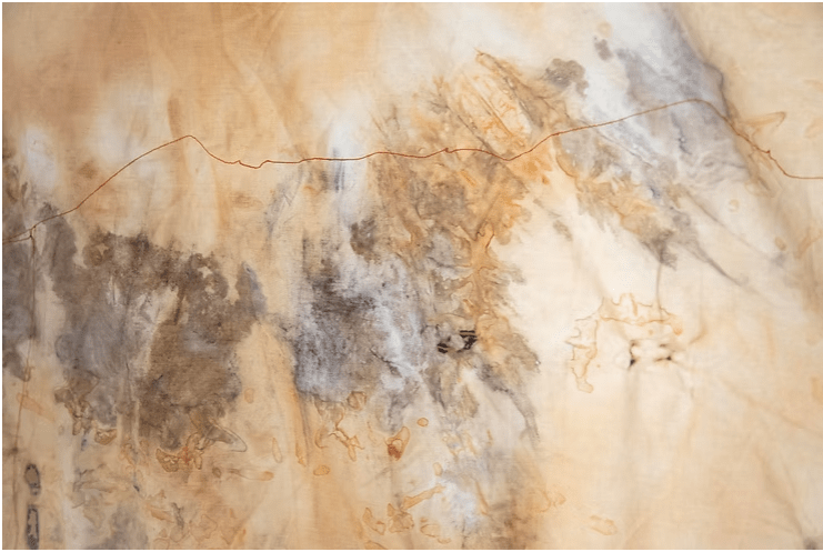 A colour photograph of a peice of cloth that is naturally dyed, rust dyed and inked, with a machine sewn red line to punctuate the landscape. The artwork and photograph are by artist Megan Kennedy