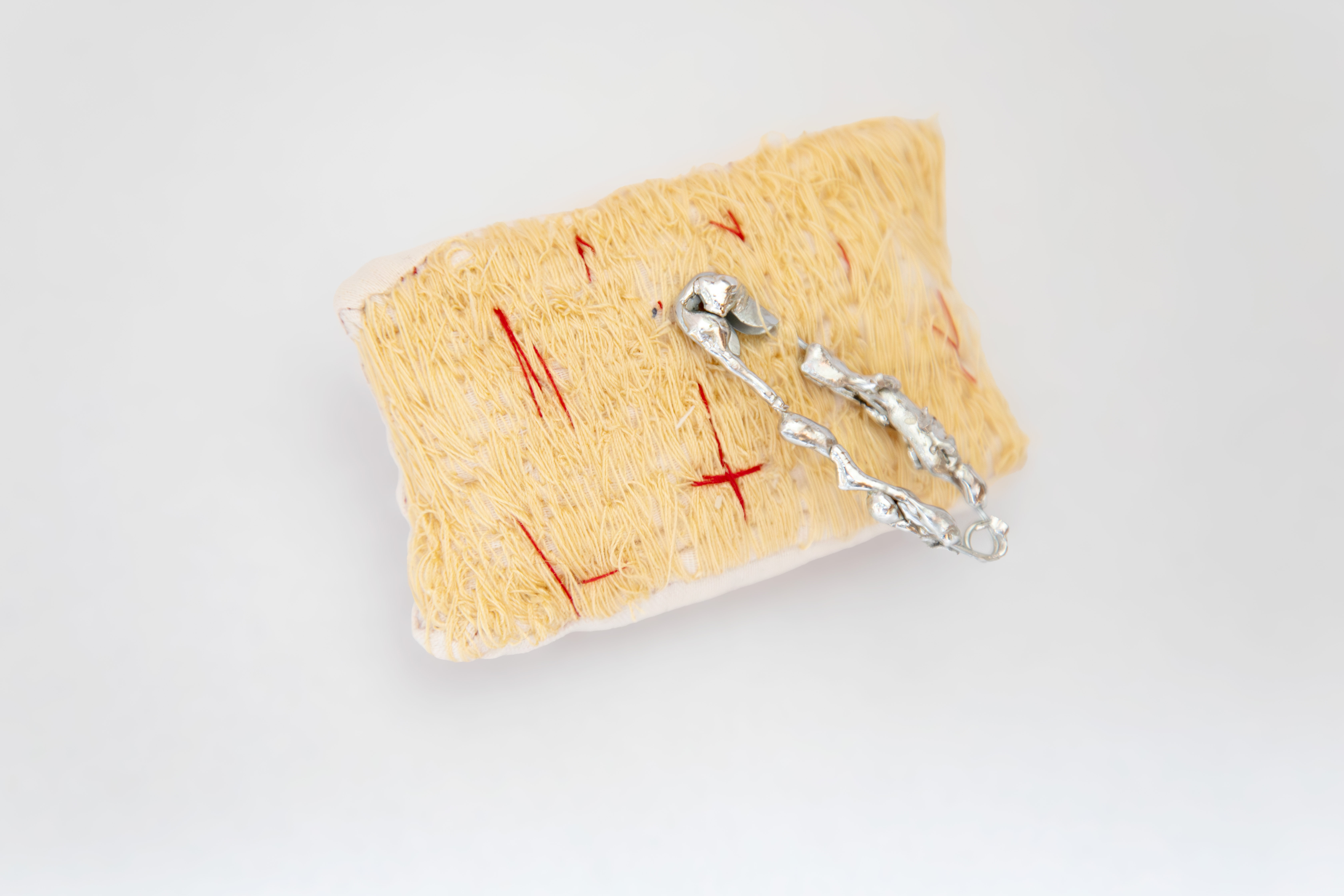 MeganKennedy,Sun,2025,Textile_and_Solder,4.5cmx7.5cm Small beige pincushion brooch with red and off-white stitching and a large, gnarled silver metal sculptural pin object