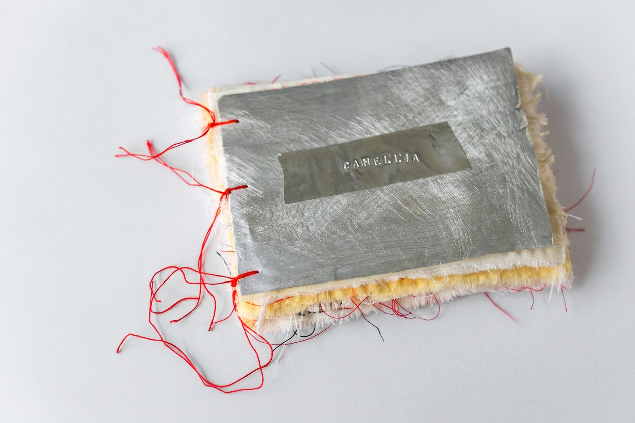 Megan_Kennedy_Camellia_Artist_Book_Front An artist book on a white minimal background made by textile artist Megan Kennedy. The book is made up of textile pages with an recycled aluminum cover embedded with the title 'Camellia' and bound with red thread.