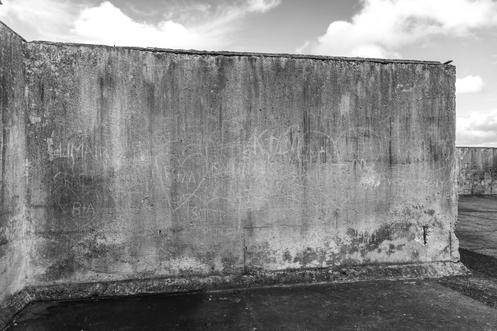Black and white photograph of a weathered concrete structure with faded graffiti, mineral staining, and worn architectural surfaces, exploring texture, materiality, and the passage of time.
