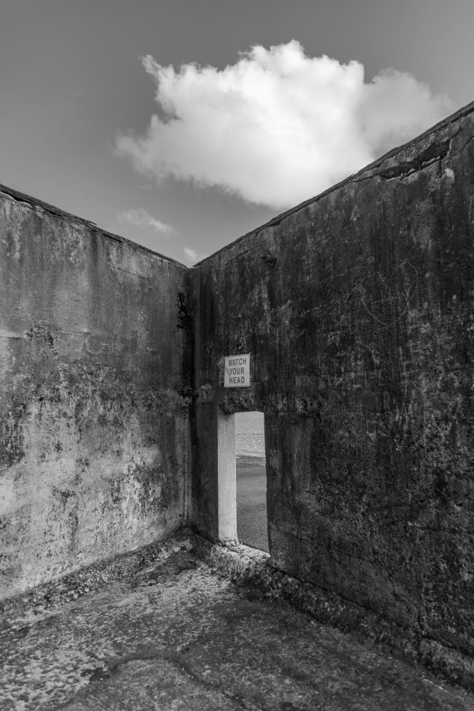 Black and white photograph of an aged concrete room with stained walls, a doorway marked "Watch Your Head," and clouds above with the sea visible beyond.