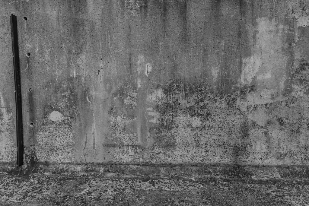 Black and white photograph of a weathered industrial concrete wall with layered mineral stains, faded markings, and a lone steel post, exploring texture, materiality, and weathering.