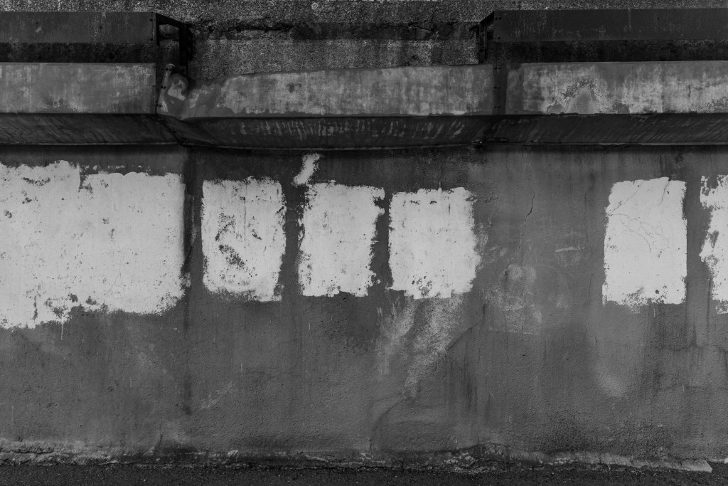 Black and white photograph of an industrial concrete wall with weathered white painted rectangles, steel beams, and stained surfaces, exploring geometry, materiality, and the passage of time. Megan Kennedy