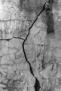 Black and white photograph of a weathered concrete wall with a prominent diagonal fissure, fine cracking, and layered mineral stains, exploring texture, materiality, and the passage of time.