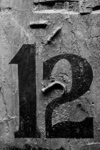Black and white photograph of a weathered industrial surface with the stencilled number 12, peeling paint, corrosion, and rich textured details.