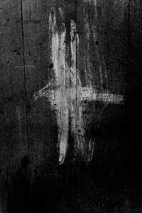 Black and white photograph of a weathered timber wall with an abstract white paint mark resembling a cross, highlighting texture, contrast, and material surface.